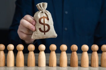 Homem segurando saco com símbolo de cifrão e alguns bonecos de madeira na frente